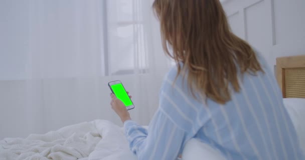 Mujer joven con teléfono celular en la cama, vista al hombro. Adicción a la tecnología. Teléfono de pantalla verde. Mujer joven tocando el vientre y el uso de smartphone en el dormitorio — Vídeos de Stock