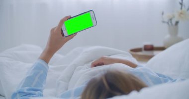 Yatakta cep telefonu olan genç bir kadın, omuz manzaralı. Teknoloji bağımlılığı. Yeşil ekran telefon. Genç bir kadın karnına dokunuyor ve yatak odasında akıllı telefon kullanıyor.