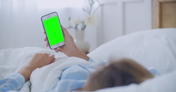Jonge vrouw met mobiele telefoon op bed, schouderzicht. Technologieverslaving. Een groene telefoon. Jonge vrouw aanraken buik en met behulp van smartphone in de slaapkamer — Stockvideo