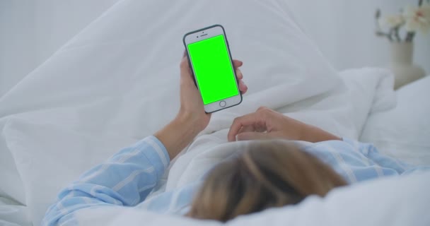 Молодая женщина с мобильным телефоном на кровати, вид на плечо. Технологическая зависимость. Зеленый экран телефона. Молодая женщина трогает живот и использует смартфон в спальне — стоковое видео