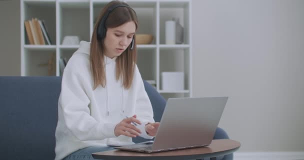 Egy fiatal nő fejhallgatós konferenciát visel, ami laptopbeszélgetésekre hív online tanárokat, tanulnak, otthonról dolgoznak. Női diák és tanulás segítségével számítógépes webkamera chat jegyzetel. Távolsági oktatás koncepciója — Stock videók
