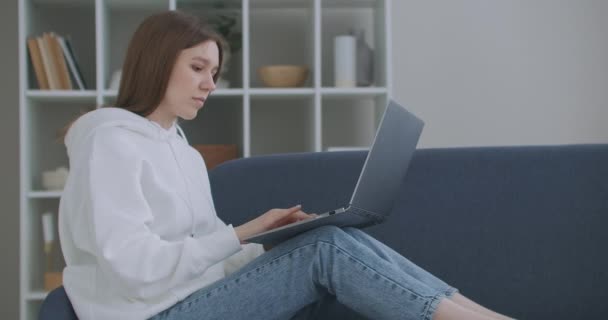 Een vrouw die thuis een laptop op de bank gebruikt. Bedachtzame jonge vrouw die met de computer op de bank zit, naar buiten kijkt, zich concentreert op het werk, zich verveelt, extra motivatie nodig heeft, op afstand thuis werkt. — Stockvideo