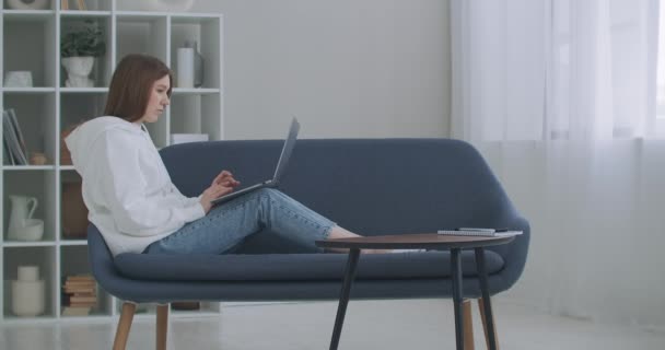 Een vrouw die thuis een laptop op de bank gebruikt. Bedachtzame jonge vrouw die met de computer op de bank zit, naar buiten kijkt, zich concentreert op het werk, zich verveelt, extra motivatie nodig heeft, op afstand thuis werkt. — Stockvideo