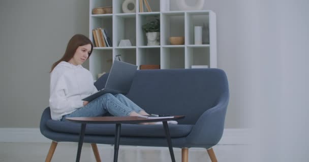 Laag zicht op een vrouw die op een bank zit en naar haar laptop kijkt. Freelance carrière. Vrolijke dame werken op laptop computer zitten op de bank thuis. — Stockvideo