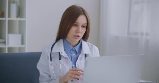 Γυναίκα επικεφαλής γιατρός του νοσοκομείου επικοινωνεί με τους γιατρούς μέσω online τηλεδιάσκεψης στο φορητό υπολογιστή, κάθεται στο γραφείο της — Αρχείο Βίντεο