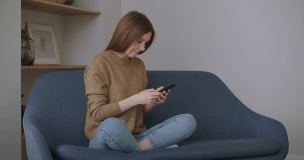 Uma mulher escrevendo uma mensagem em seu novo smartphone enquanto se senta no sofá com um travesseiro branco em casa. — Vídeo de Stock