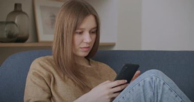 Evdeki ofiste masa başında oturan genç bir kadın, elinde akıllı bir telefonla elinde telefon tutarak arkadaşlarıyla sohbet ederken modern cihazlar ve internet bağlantısı kullanıyor.