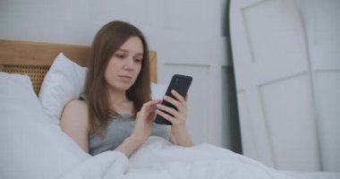 Sabahları yatak odasında oturan kadın yatağın yumuşak derisine yaslanıyor. Akıllı telefonunuzla mesajlar yazın, bilgi almak için uygulamaları kullanın