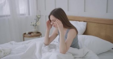 Yatakta başını tutarak oturan kadınlar. Onun acı verici bir baş ağrısı var. Kadın baş ağrısı çekiyor, şakaklarını ovuyor ve acı içinde kaşlarını çatıyor. Mevsimlik hastalıklar, yatak odasındaki beyaz kız hastalık izninde.