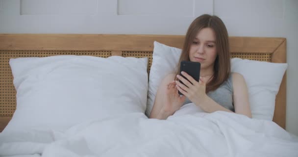 Face Close-up na mulher digitando uma mensagem em um telefone celular usando 2 mãos enquanto confortavelmente deitado em uma cama em um quarto de hotel. mulher usando telefone inteligente e deitado na cama de manhã depois de acordar — Vídeo de Stock