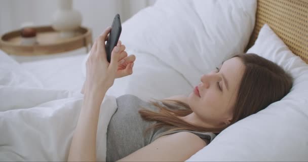 Face Close-up na mulher digitando uma mensagem em um telefone celular usando 2 mãos enquanto confortavelmente deitado em uma cama em um quarto de hotel. mulher usando telefone inteligente e deitado na cama de manhã depois de acordar — Vídeo de Stock