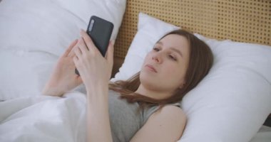 Sabahları yatak odasında oturan kadın yatağın yumuşak derisine yaslanıyor. Akıllı telefonunuzla mesajlar yazın, bilgi almak için uygulamaları kullanın