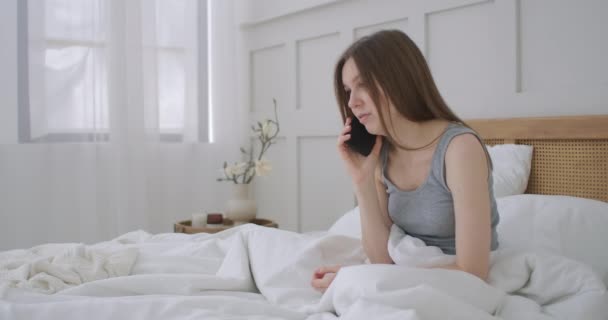 Una joven caucásica sentada en la cama por la mañana se siente mal. Una joven está haciendo una llamada de teléfono inteligente a su médico. — Vídeo de stock