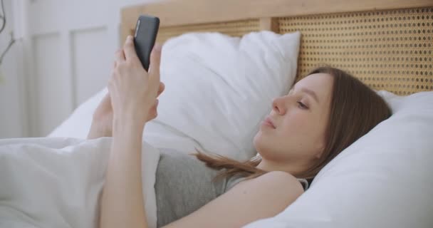 Лицо крупным планом женщины, печатающей сообщение на мобильном телефоне двумя руками, удобно лежащей на кровати в гостиничном номере. женщина с помощью смартфона и лежа на кровати утром после пробуждения — стоковое видео