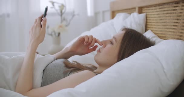 Femme le matin assise dans la chambre appuyée contre le dos en cuir souple du lit. Écrire des messages avec votre smartphone, utiliser des applications pour l'information — Video