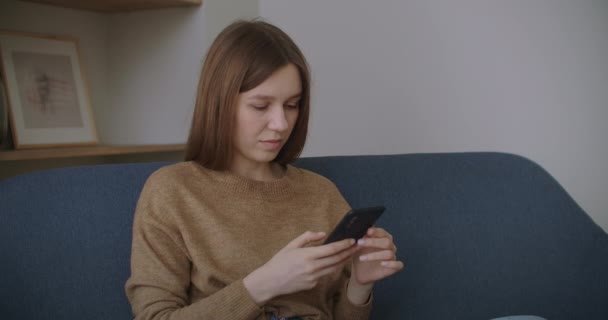 Jovem mulher sentada na mesa no escritório em casa distraída do trabalho segurando telefone inteligente usando o site de redes sociais conversando com o amigo remotamente usando dispositivo moderno e conexão com a internet — Vídeo de Stock