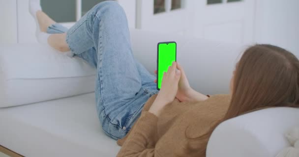 Mulher em casa deitada em um sofá usando Smartphone com tela de Mock-up verde, fazendo Swiping, Scrolling Gestures. Um tipo a usar telemóvel, a navegar nas redes sociais da Internet. Ponto de vista Camera Shot — Vídeo de Stock