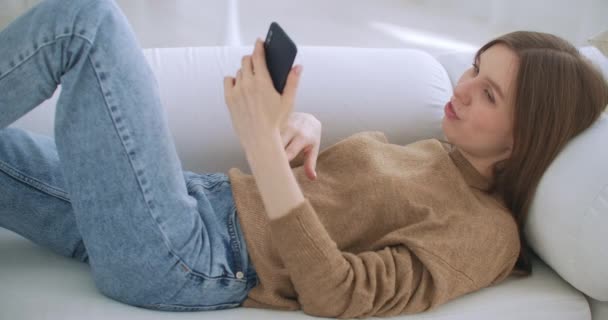 Жінка лежить на дивані у вітальні, спілкуючись, пишучи повідомлення за допомогою смартфона. Самотня щаслива леді розслабляється за допомогою смартфона, що сидить на дивані читання, написання, пошук, перегляд на мобільному Інтернеті — стокове відео