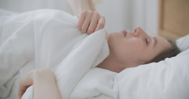 Kranke Frau schläft im Bett unter Decke. Frau bedeckt ihre Ohren mit einer Bettdecke. Junge Frau versteckt sich unter Decke auf einem Bett. — Stockvideo