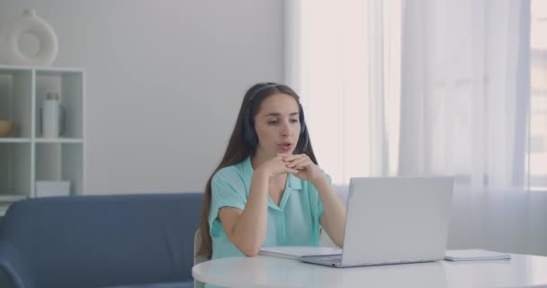 Estudante mulher sentar-se na mesa em casa usar fone de ouvido falar distantemente por aplicativo de chamada de vídeo e dispositivo laptop e-aprender idioma estudando com e tutor ou colega de classe fazer tarefa comum remotamente, conceito de tecnologia moderna — Vídeo de Stock