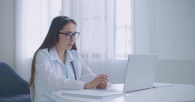 Kadın tıp asistanı beyaz önlük giyiyor, kulaklık takıyor uzaktan hastayı laptoptan arıyor. Doktor sanal sohbet bilgisayar uygulaması kullanarak müşteriyle konuşuyor. Teletıp, uzaktan sağlık hizmeti kavramı.