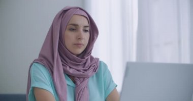 Güzel genç Müslüman kadın iş yerinde dizüstü bilgisayarı üzerinde çalışıyor. Genç Müslüman bir kadın, dizüstü bilgisayarın önünde oturuyor, kafasını kaldırıp web kamerasına bakıyor.