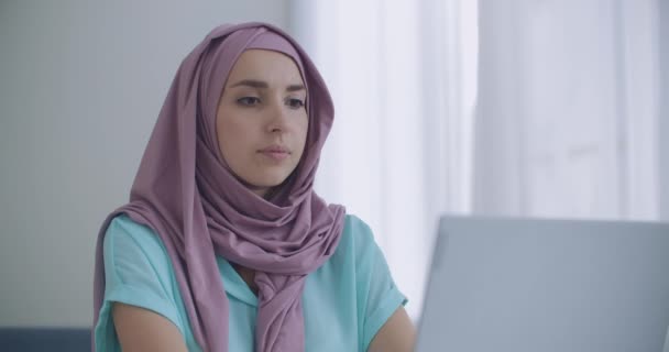 Wanita muslim bekerja pada laptop di kantor dengan rekan kerja Kristennya. Potret perempuan muslim muda dalam jilbab menggunakan laptop di dalam ruangan apartemen — Stok Video