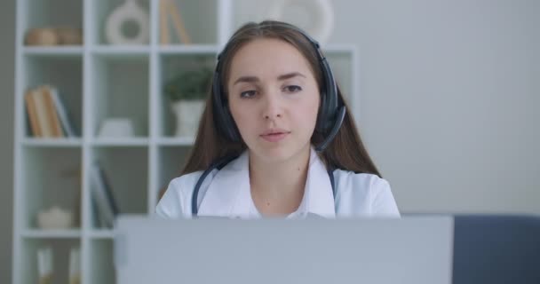 Zdravotnice nosí bílý kabát, sluchátka video volá vzdáleného pacienta na notebook. Doktor mluví s klientem pomocí virtuální chatovací aplikace. Telemedicína, koncept vzdálených zdravotnických služeb. — Stock video