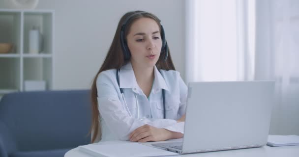 Asistente médica femenina lleva abrigo blanco, auriculares de vídeo llamando a paciente distante en el ordenador portátil. Doctor hablando con el cliente utilizando la aplicación de chat virtual de la computadora. Telemedicina, concepto de servicios sanitarios remotos. — Vídeos de Stock