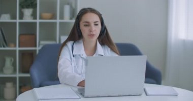 Kadın tıp asistanı beyaz önlük giyiyor, kulaklık takıyor uzaktan hastayı laptoptan arıyor. Doktor sanal sohbet bilgisayar uygulaması kullanarak müşteriyle konuşuyor. Teletıp, uzaktan sağlık hizmeti kavramı.