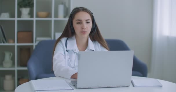 Kvinnlig medicinsk assistent bär vit rock, headset video ringer avlägsen patient på bärbar dator. Läkare pratar med klienten med hjälp av virtuell chatt dator app. Telemedicin, distansvård. — Stockvideo