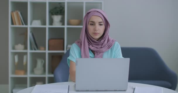 Schöne junge Muslimin arbeitet an ihrem Arbeitsplatz am Laptop. Eine junge Muslimin sitzt vor einem Laptop-Bildschirm, blickt auf und blickt in die Webkamera — Stockvideo