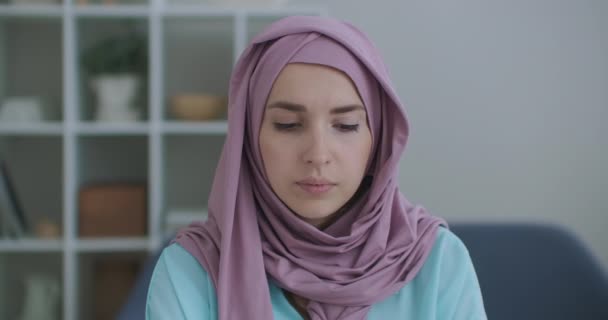 Mooie jonge moslim vrouw werkt aan laptop op haar werkplek. Een jonge moslim vrouw, zittend voor een laptop scherm, kijkt op en kijkt naar de webcamera — Stockvideo
