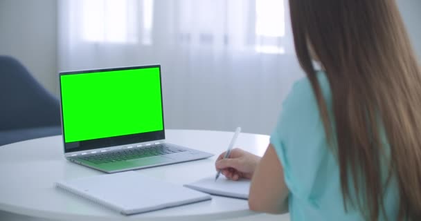 La mujer se sienta en el escritorio en el dormitorio, mira la pantalla verde del ordenador portátil y habla con alguien a través de las comunicaciones de vídeo de Internet, a veces tomando notas en el cuaderno. Primer plano — Vídeos de Stock