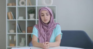 Başörtülü Müslüman bir kadın masaya oturur ve elini sallayarak kameraya bakar ve konuşur. Vlog ya da video tesettürlü bir kadını çağırıyor. Konuşurken Arap bir kadının kafasının görüntüsü.