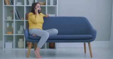 Oturma odasında modern bir şekilde otururken akıllı telefonuyla konuşan mutlu olgun bir kadın. Teknoloji ve rahatlama kavramı.