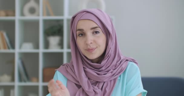 A Muslim woman in a hijab sits at a table and looks into the camera waving her hand and talking. vlog or video call a woman in a hijab. shot of the head of an Arab woman in conversation — Stock Video