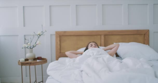 Schöne Brünette wacht am Morgen langsam auf, dehnt sich und steht aus dem Bett auf, die Sonne scheint aus dem großen Fenster auf sie. Glückliches junges Mädchen begrüßt neuen Tag — Stockvideo