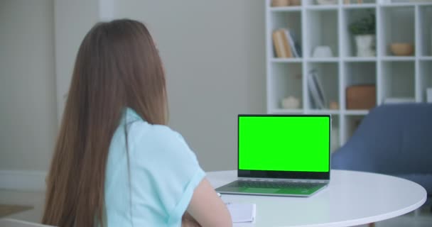 La mujer se sienta en el escritorio en el dormitorio, mira la pantalla verde del ordenador portátil y habla con alguien a través de las comunicaciones de vídeo de Internet, a veces tomando notas en el cuaderno. Primer plano — Vídeos de Stock