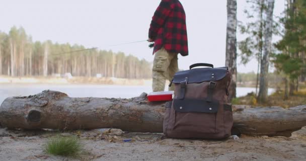 Człowiek wędkuje w jeziorze, stoi na wybrzeżu w lesie, plecak ze sprzętem i przynętami na ziemi, relaksuje się hobby — Wideo stockowe