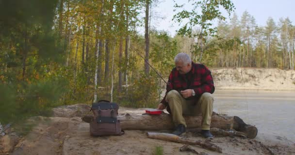 Laki-laki paruh baya membuka kotak dengan umpan memancing dan umpan duduk di log di tepi sungai, mempersiapkan diri untuk memancing — Stok Video