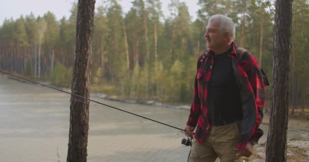 Fisherman is dressed red checkered shirt and carrying spin fishing rod, walking on high coast of lake — Stock Video