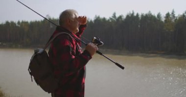 Turist, güneşli bir sonbahar gününde ormanda nehir veya göl manzarası seyrediyor. Yüzü avuçla avuçla kaplanmış, gölün kıyısında balıkçı var.