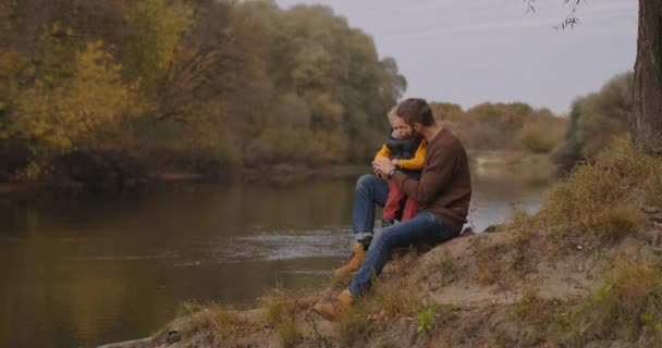Felice padre sta comunicando con il suo piccolo figlio in natura, uomo e ragazzo sono seduti sulla costa del fiume foresta insieme nel fine settimana — Video Stock