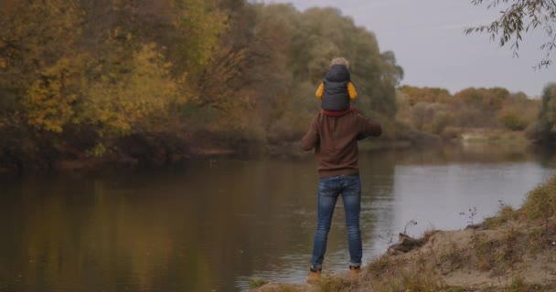 Vater und kleiner Sohn am Ufer des Flusses am Herbsttag, Kleinkind sitzt auf den Schultern von Papa, Rückansicht, Familienwochenende in der Natur — Stockvideo