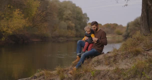 Vater und kleiner Sohn verbringen Zeit zusammen am Ufer des kleinen Waldflusses, ruhen sich im Herbst in der Natur aus, sind glückliche Eltern und Kinder — Stockvideo