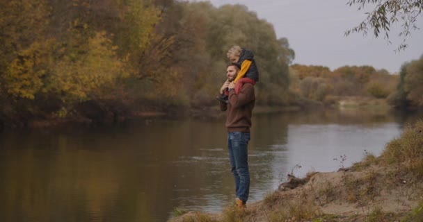 Joyful man is holding child boy on shoulders and enjoying nature of river and forest at autumn, happy father and son at weekend walk — Stock Video