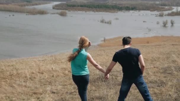 Молодая пара проходит через холм у реки держа за руки задний вид — стоковое видео