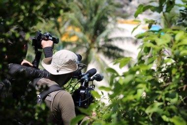 Photographer or videographer holding the video camera on a tripod on top of the mountain. Forest,ocean and blue sky  background.