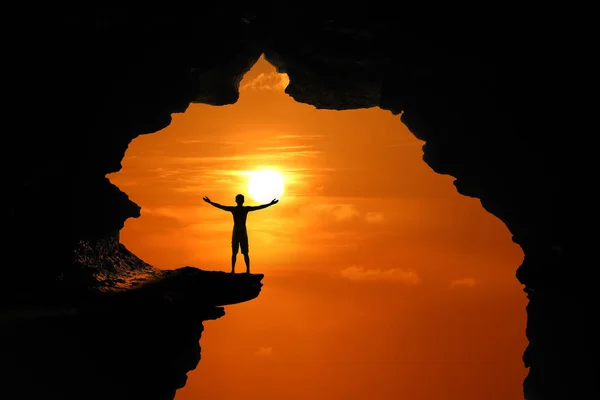 El hombre de pie en una cueva en un alto acantilado al atardecer del cielo rojo —  Fotos de Stock